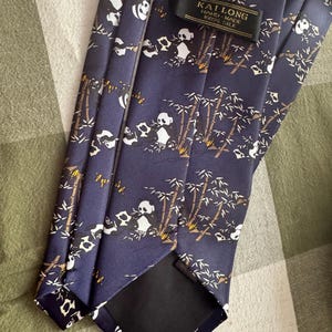 May include: A dark blue necktie featuring a repeating pattern of black and white pandas eating bamboo stalks. The tie includes a black fabric label with the text KAI LONG HAND-MADE 100% SILK stitched near the top of the garment.