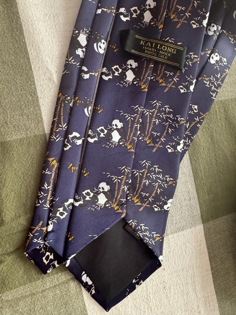 May include: A dark blue necktie featuring a repeating pattern of black and white pandas eating bamboo stalks. The tie includes a black fabric label with the text KAI LONG HAND-MADE 100% SILK stitched near the top of the garment.