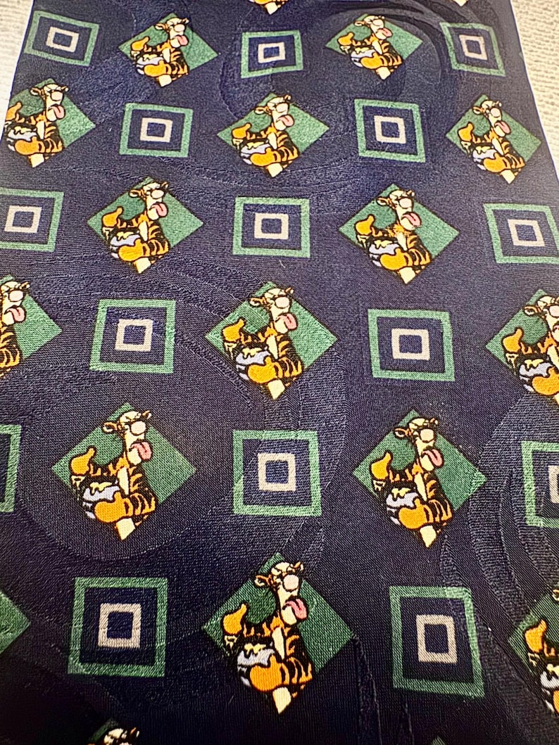 K&ouml;nnte beinhalten: Marineblaue Krawatte mit einem sich wiederholenden Muster der Figur Tigger aus Winnie Puuh. Das Design umfasst Tigger in einer Raute, umgeben von gr&uuml;nen Rauten und blauen Quadraten. Die Krawatte ist aus glattem Material, m&ouml;glicherweise Seide.