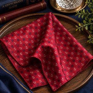 May include: A square pocket square in a deep red color featuring a repeating pattern of small white anchors and dark blue ship wheels. The fabric has a smooth, slightly reflective finish and is folded to display the nautical-themed print clearly.