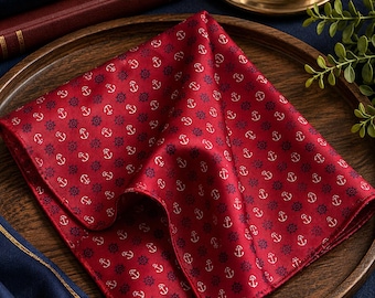 Puede incluir: Un pañuelo de bolsillo cuadrado de color rojo intenso con un patrón repetitivo de pequeñas anclas blancas y timones de barco azul oscuro. La tela tiene un acabado liso y ligeramente brillante, doblado para mostrar el estampado náutico.