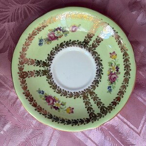 May include: A pale green porcelain saucer featuring a central white circular area, decorated with gold leaf laurel wreath patterns and small clusters of colorful floral motifs including pink roses, yellow flowers, and blue blossoms.