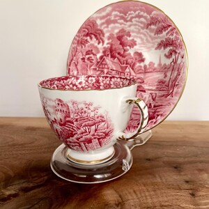 Pu&ograve; includere: Un set di tazza e piattino bianchi con un disegno di paesaggio rosso. La tazza ha un manico e un bordo dorati, e il piattino ha un bordo dorato abbinato. Il set &egrave; esposto su un supporto trasparente.