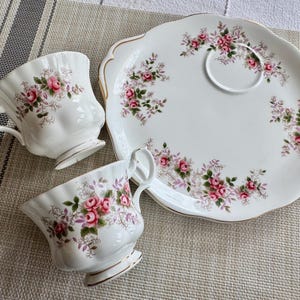 May include: A white teacup and saucer set with a floral pattern featuring pink roses and green leaves. The set is on a white and gray patterned surface.