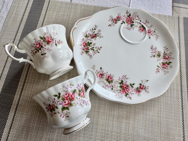 May include: A white teacup and saucer set with a floral pattern featuring pink roses and green leaves. The set is on a white and gray patterned surface.