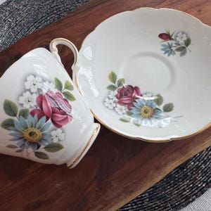 May include: A white teacup and saucer set with a floral design. The cup and saucer feature a delicate floral pattern with pink, blue, and white flowers. The saucer has a gold trim.