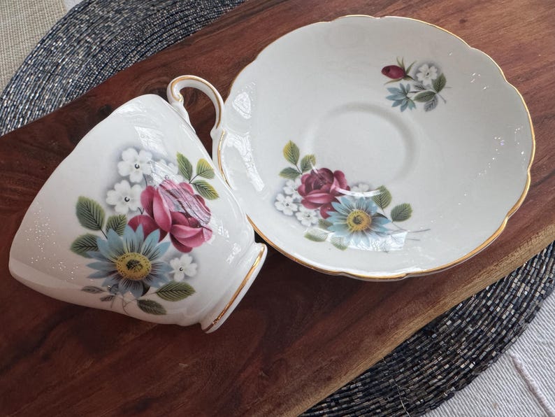 May include: A white teacup and saucer set with a floral design. The cup and saucer feature a delicate floral pattern with pink, blue, and white flowers. The saucer has a gold trim.