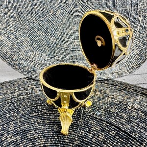 May include: A black and gold egg-shaped trinket box with a hinged lid and three gold feet. The box is open and the inside is lined with black velvet.