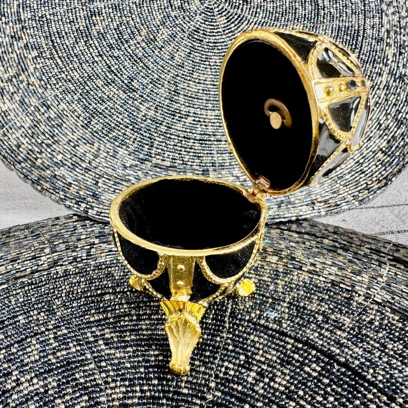 May include: A black and gold egg-shaped trinket box with a hinged lid and three gold feet. The box is open and the inside is lined with black velvet.