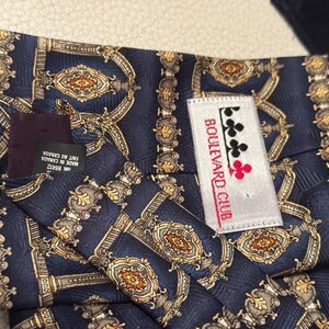 May include: A navy blue necktie featuring a repeating gold and silver ornate architectural pattern. A white label is stitched onto the back with the text Boulevard Club and a clover icon. A black tag reads Made in Canada and RN 85412.
