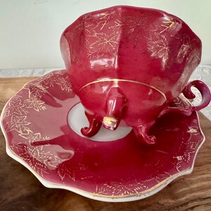 May include: A burgundy teacup and saucer set with gold leaf accents. The teacup has a curved handle and sits on a matching saucer. The set is made of porcelain and has a scalloped edge.