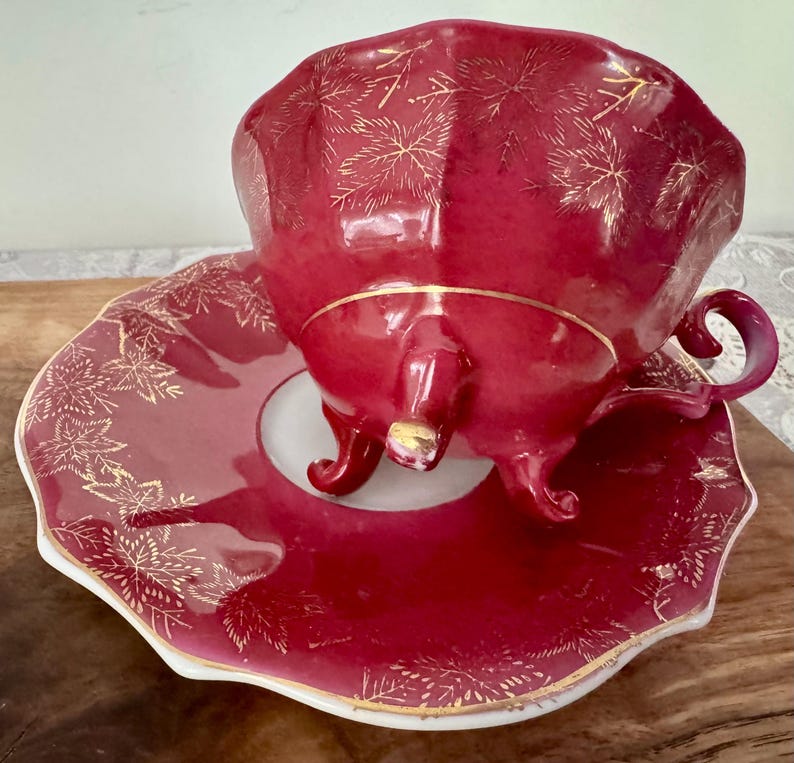 May include: A burgundy teacup and saucer set with gold leaf accents. The teacup has a curved handle and sits on a matching saucer. The set is made of porcelain and has a scalloped edge.