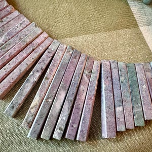 May include: A necklace featuring a series of rectangular, flat stone beads in shades of dusty pink, mauve, and muted green. The beads are arranged in a fan-like pattern, with additional round green stone beads visible at the top right corner.