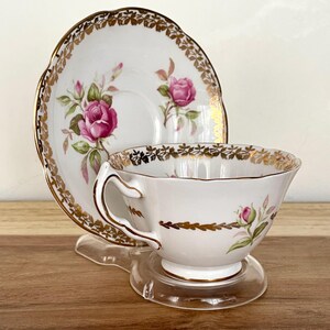 Puede incluir: Juego de taza y platillo de t&eacute; blanco con dise&ntilde;o de rosas. La taza y el platillo tienen ribetes dorados y un patr&oacute;n de rosas rosas. La taza est&aacute; sobre un soporte de pl&aacute;stico transparente, y el platillo est&aacute; detr&aacute;s.