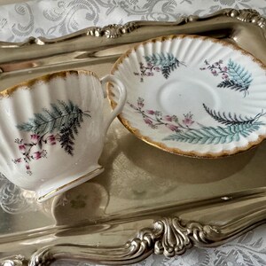 Puede incluir: Juego de taza y platillo blancos con borde dorado y dise&ntilde;o de helechos y flores. La taza y el platillo est&aacute;n colocados en una bandeja plateada. La taza tiene un dise&ntilde;o acanalado.