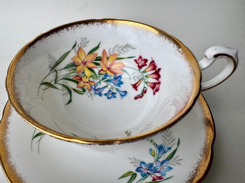May include: A white teacup and saucer with a gold rim and floral pattern. The flowers are pink, yellow, and blue.