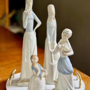 K&ouml;nnte beinhalten: Eine Sammlung von Porzellanfiguren, die Frauen in langen Kleidern darstellen. Die Figuren sind &uuml;berwiegend wei&szlig; mit hellblauen Akzenten. Eine Figur h&auml;lt einen Korb mit einem Vogel, eine andere ein kleines Kind. Die Figuren stehen auf einem Marmortablett.