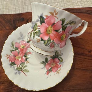 Puede incluir: Un juego de taza y platillo de t&eacute; blanco con un dise&ntilde;o floral con rosas rosas y hojas verdes. El juego tiene un borde dorado.