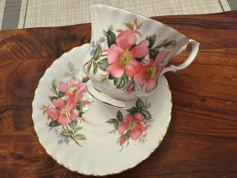 Puede incluir: Un juego de taza y platillo de t&eacute; blanco con un dise&ntilde;o floral con rosas rosas y hojas verdes. El juego tiene un borde dorado.