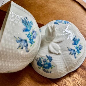 May include: A white porcelain trinket box with a lid, featuring a blue bird and floral design. The box is open, revealing its interior. The lid has a rose-shaped handle. The box is on a wooden surface.