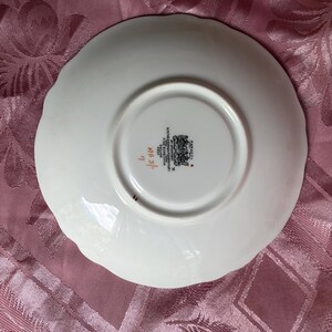 May include: The underside of a white bone china saucer with a scalloped edge. The center features a black stamp reading Paragon, By Appointment, H.M. The Queen & H.M. Queen Mary, Fine Bone China, England, Regd, with the handwritten red text A53/7.