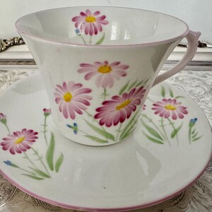 May include: White teacup and saucer set with a floral daisy design in pink and yellow. The cup and saucer have a pink trim. The daisies are painted with green stems and leaves. The set is on a silver tray.