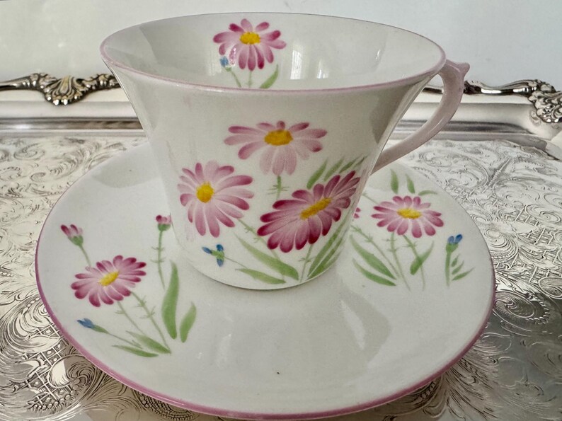 May include: White teacup and saucer set with a floral daisy design in pink and yellow. The cup and saucer have a pink trim. The daisies are painted with green stems and leaves. The set is on a silver tray.