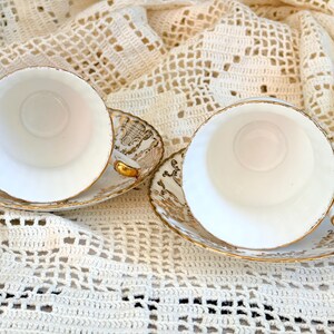 May include: Two white porcelain teacups and matching saucers featuring intricate gold filigree patterns along the rims and edges. The cups have smooth white interiors and gold-trimmed handles, while the saucers display detailed gold scrollwork.