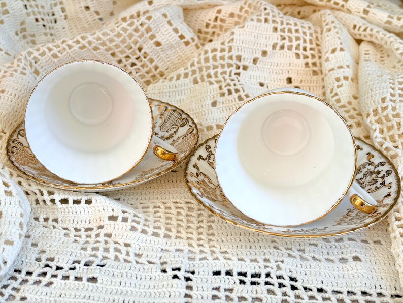 May include: Two white porcelain teacups and matching saucers featuring intricate gold filigree patterns along the rims and edges. The cups have smooth white interiors and gold-trimmed handles, while the saucers display detailed gold scrollwork.