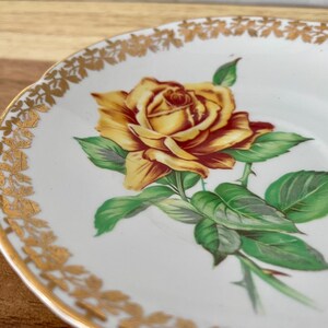 Puede incluir: Platillo de cer&aacute;mica blanca con un borde floral dorado y una ilustraci&oacute;n de rosa amarilla y roja. La rosa tiene hojas verdes y espinas. El platillo est&aacute; sobre una superficie de madera.