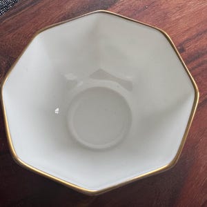 Pode incluir: Uma tigela branca octogonal com borda dourada. A tigela tem um design simples e elegante e provavelmente &eacute; feita de cer&acirc;mica ou porcelana. A tigela est&aacute; vazia e repousa sobre uma superf&iacute;cie de madeira escura.