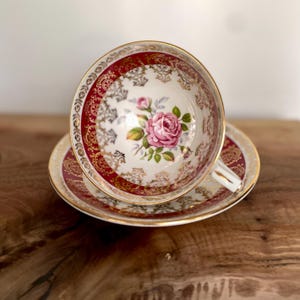 Pode incluir: Um conjunto de ch&aacute; vintage com ch&aacute;vena e pires. A ch&aacute;vena e o pires apresentam um fundo branco com um desenho floral de rosas cor-de-rosa. As bordas s&atilde;o adornadas com ouro e um padr&atilde;o bord&ocirc;. A ch&aacute;vena de ch&aacute; est&aacute; sobre o pires.