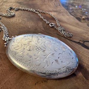 Puede incluir: Collar con colgante ovalado de plata con intrincado grabado floral. El colgante cuelga de una delicada cadena. La superficie del colgante est&aacute; muy pulida, reflejando la luz. El dise&ntilde;o presenta un patr&oacute;n detallado de flores y hojas.