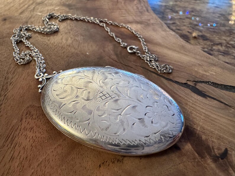 Puede incluir: Collar con colgante ovalado de plata con intrincado grabado floral. El colgante cuelga de una delicada cadena. La superficie del colgante est&aacute; muy pulida, reflejando la luz. El dise&ntilde;o presenta un patr&oacute;n detallado de flores y hojas.