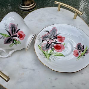 Puede incluir: Juego de taza y platillo blancos con un dise&ntilde;o floral de flores rojas, negras y rosas y hojas verdes. La taza est&aacute; sobre una bandeja de m&aacute;rmol blanco con asas doradas. El platillo tiene un borde dorado.