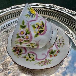 May include: A white teacup and saucer set with a floral design and pink ribbon accents. The teacup and saucer are trimmed with gold and sit on a silver tray. The teacup has a handle and is tilted.