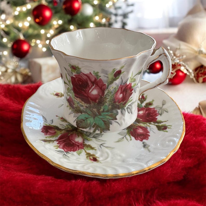 May include: A white teacup and saucer with a floral design featuring red roses and green leaves. The cup and saucer have a gold trim.