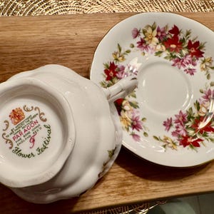 May include: A white teacup and saucer set with a floral pattern. The saucer has a gold rim and the teacup has a floral design with a gold trim. The bottom of the teacup has the words "Paragon Fine Bone China England  Regd No.  Flower Festival" printed on it.