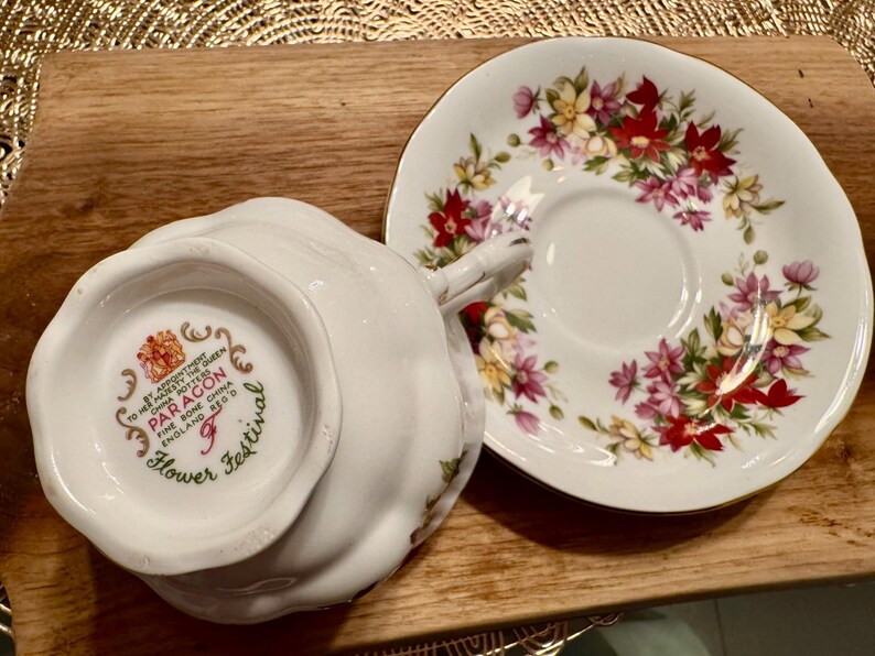 May include: A white teacup and saucer set with a floral pattern. The saucer has a gold rim and the teacup has a floral design with a gold trim. The bottom of the teacup has the words "Paragon Fine Bone China England  Regd No.  Flower Festival" printed on it.
