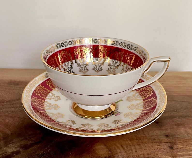 Pode incluir: Conjunto de ch&aacute; vintage branco com design vermelho e dourado. A ch&aacute;vena tem uma pega dourada e assenta num pires a condizer. O conjunto apresenta um design ornamental, ideal para a hora do ch&aacute;.