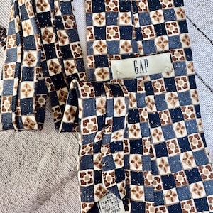 May include: A necktie featuring a checkered pattern in navy blue, light blue, and tan with a repeating diamond motif. The tie includes a white label with the text GAP and a smaller tag with the text FABRIQUE AUX ETATS-UNIS AVEC TISSU IMPORTE CA 17897.