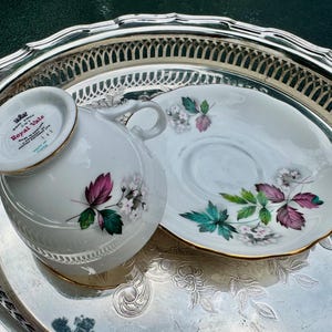 May include: A white teacup and saucer set with a floral design featuring pink, green, and white flowers, and gold trim. The teacup is upside down on a silver tray. The set is marked "Royal Vale".