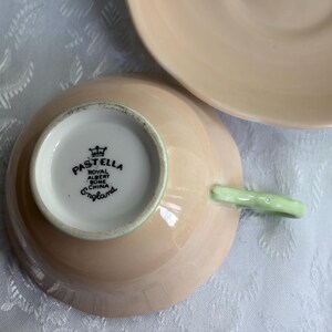 May include: A pale peach-colored bone china teacup and matching saucer. The underside of the teacup features a black stamp reading PASTELLA ROYAL ALBERT BONE CHINA ENGLAND with a crown icon. The teacup has a light green handle.