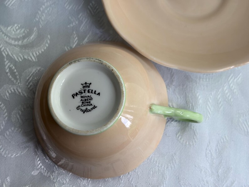 May include: A pale peach-colored bone china teacup and matching saucer. The underside of the teacup features a black stamp reading PASTELLA ROYAL ALBERT BONE CHINA ENGLAND with a crown icon. The teacup has a light green handle.
