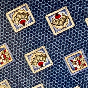 May include: A blue necktie featuring a repeating pattern of small, square-framed motifs. Each square contains a stylized, fan-like design in shades of cream, light blue, and red, set against a textured, dark blue honeycomb-like background.