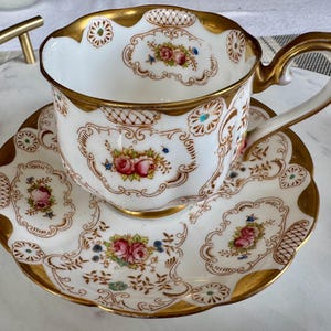 K&ouml;nnte beinhalten: Ein wei&szlig;es Tee-Set mit goldenen Verzierungen und floralen Mustern. Die Tasse und Untertasse haben einen gewellten Rand und detaillierte Designs mit rosa und blauen Blumen. Der Henkel der Tasse ist ebenfalls golden.