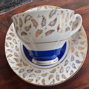 May include: A white teacup and saucer with a blue band and gold leaf pattern. The teacup has a handle and the saucer has a scalloped edge.