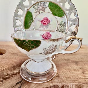 Puede incluir: Un juego de taza y platillo de t&eacute; vintage. La taza y el platillo de porcelana blanca presentan adornos dorados, una rosa rosa y detalles verdes. El platillo tiene un borde en forma de coraz&oacute;n. El juego se exhibe sobre una superficie de madera.