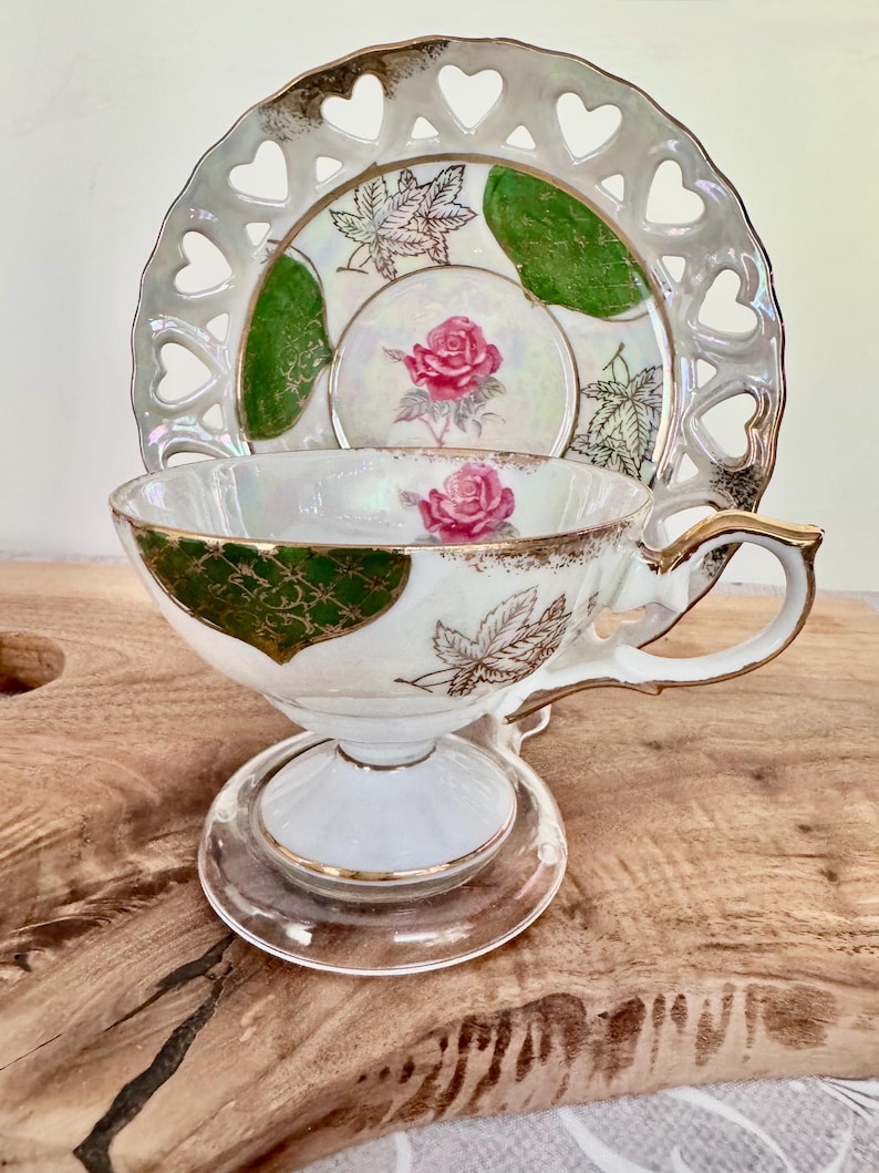 Puede incluir: Un juego de taza y platillo de t&eacute; vintage. La taza y el platillo de porcelana blanca presentan adornos dorados, una rosa rosa y detalles verdes. El platillo tiene un borde en forma de coraz&oacute;n. El juego se exhibe sobre una superficie de madera.