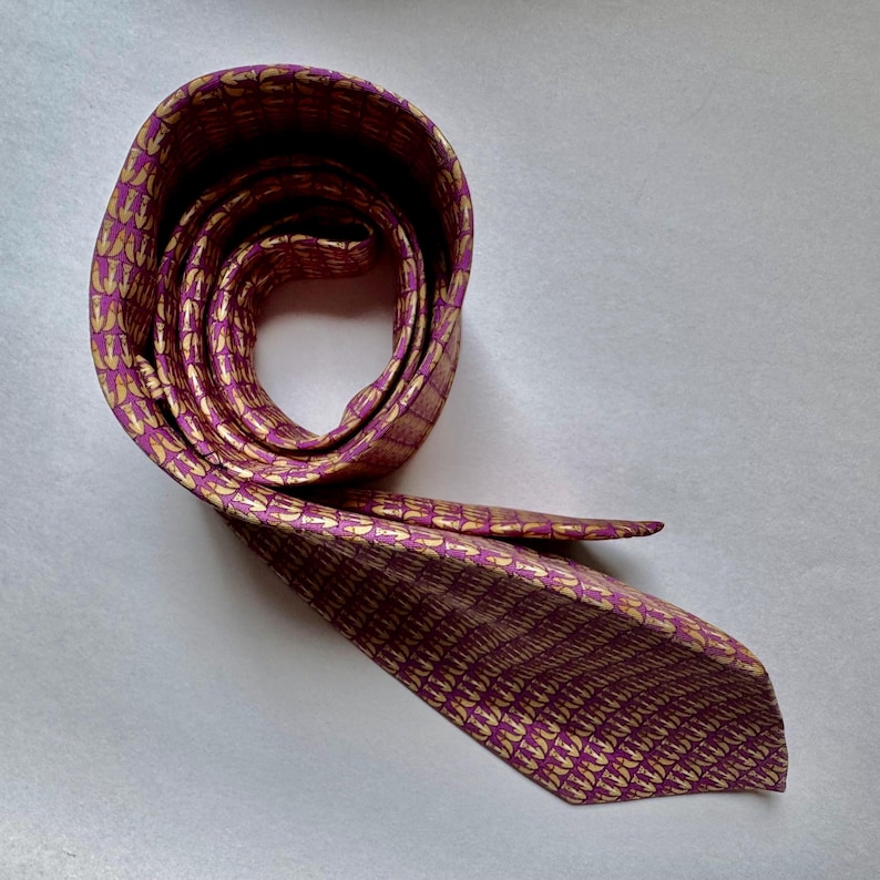 Puede incluir: Una corbata con un fondo magenta vibrante y un patr&oacute;n repetitivo de peque&ntilde;os motivos en forma de flecha de color amarillo dorado. La corbata est&aacute; enrollada en forma circular, con un extremo extendido hacia afuera mostrando la punta.